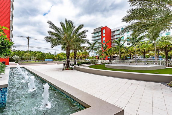View of a modern multi-story building with a red facade, surrounded by palm trees and water features.
