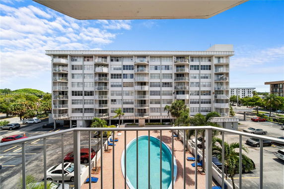 View from a balcony overlooking a multi-story residential building with balconies. A swimming pool is visible below, surrounded by a patio area and some palm trees. Cars are parked in a parking area around the building.