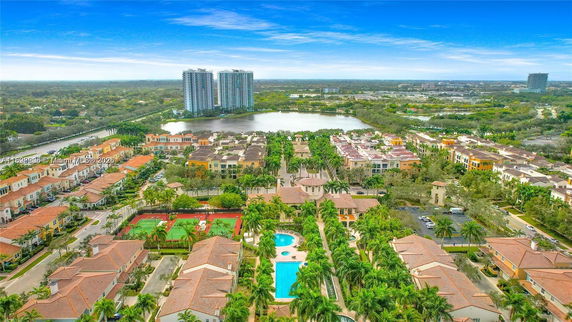 Wide-angle view of a residential area with multiple buildings, pool, and tennis courts, surrounded by greenery.
