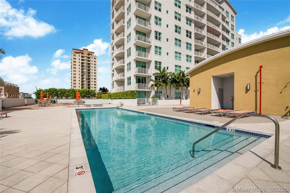 Outdoor swimming pool adjacent to a multi-story building with lounge chairs and umbrellas.