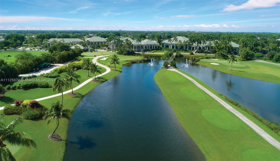 Aerial view of a large estate with multiple buildings surrounded by manicured lawns, a golf course, a pond, and water fountains.