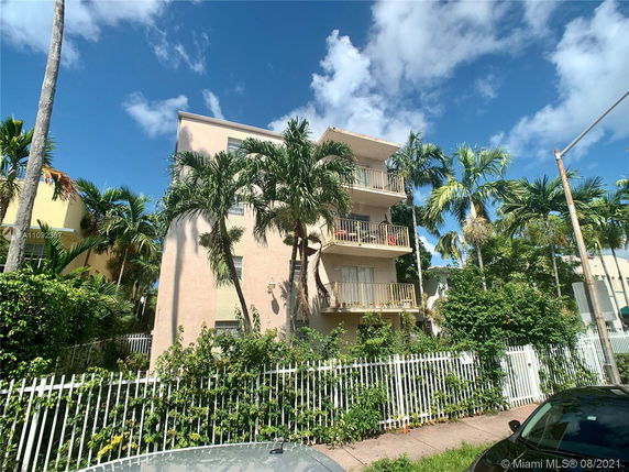 Front view of a three-story building with balconies surrounded by palm trees.