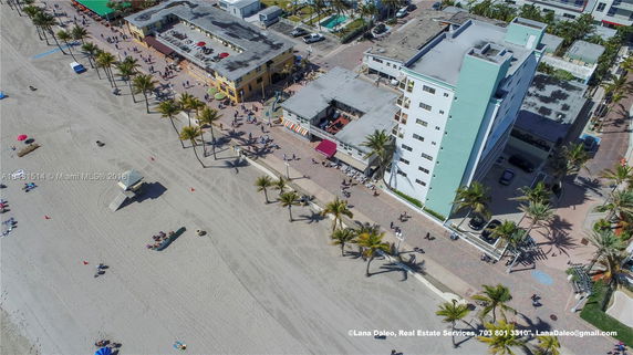 Aerial view of beachfront buildings with flat rooftops, surrounded by a beach and palm trees.
