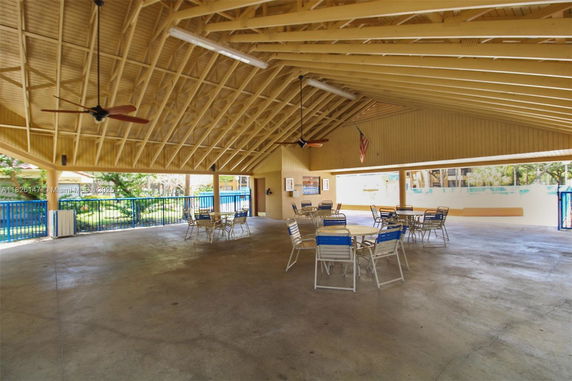 Spacious open clubhouse area with tables and chairs under a high ceiling, featuring fans and natural lighting.