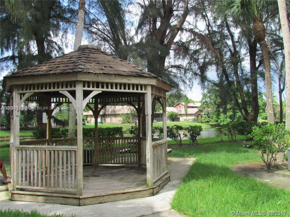 Wooden gazebo in a lush garden setting with trees and a pond in the background.