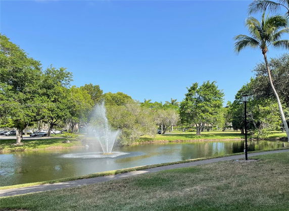 View of a park with a pond and a water fountain, surrounded by trees and a pathway.