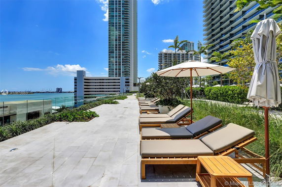 Rooftop area with lounge chairs and umbrellas, overlooking city skyscrapers and a body of water.
