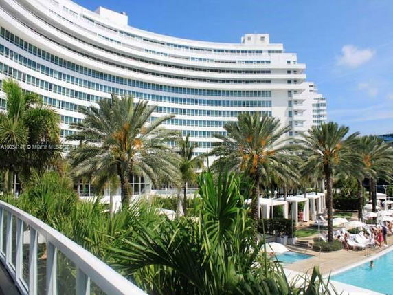 Curved multi-story hotel building with numerous windows and palm trees in front, near a swimming pool area.