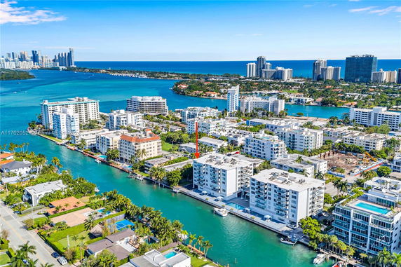 Aerial view of a coastal cityscape with multiple multi-story residential buildings lined beside a waterway, surrounded by palm trees and other urban structures.