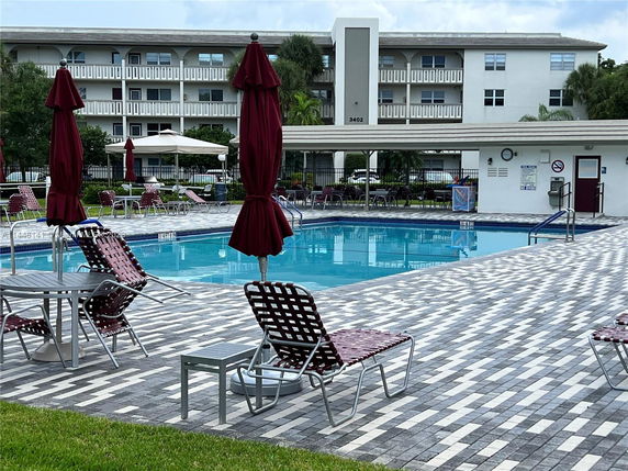 Open area with a swimming pool surrounded by lounge chairs and parasols, adjacent to a multi-story residential building.