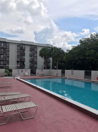 Outdoor swimming pool area with lounge chairs and a multi-story building in the background.