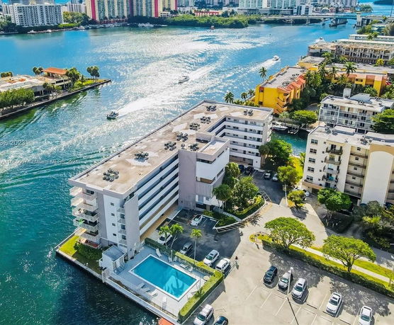 Aerial view of a multi-story building near a waterfront with surrounding buildings and a parking area.