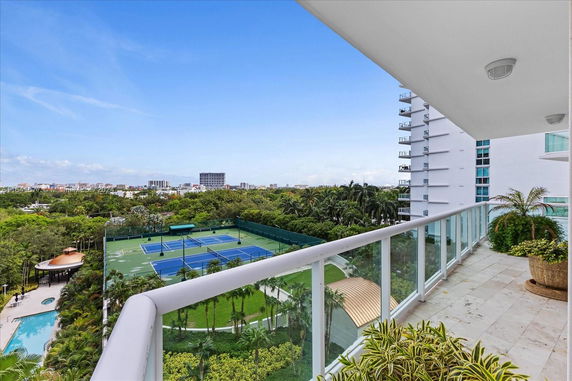 View from a balcony overlooking tennis courts, a swimming pool, and distant city buildings.