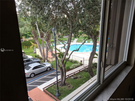 View from a window showing a parking area and a swimming pool surrounded by trees.