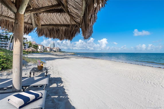 View from under a beach hut looking out at a sandy beach, ocean, and nearby buildings.
