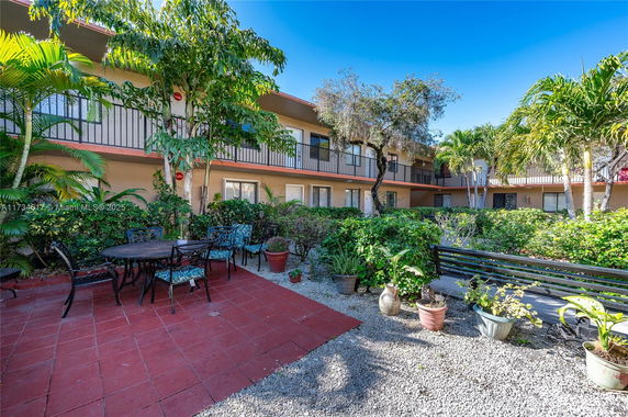 Outdoor seating area with chairs and tables surrounded by greenery and a view of a two-story building with balconies.