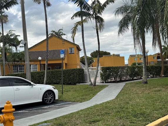 View of yellow-colored buildings with tiled roofs and a fenced-in area. Palm trees and parked cars are visible in the foreground.
