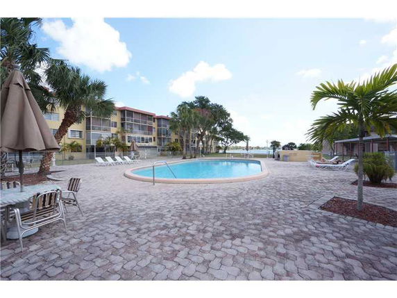 Outdoor area with a swimming pool surrounded by lounge chairs and tables, adjacent to a multi-story building.
