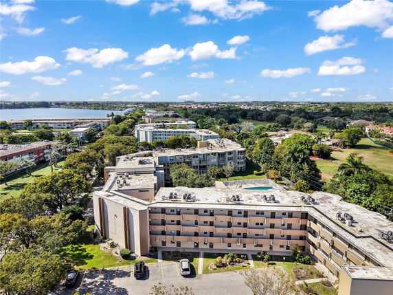 Aerial view of a multi-story building with flat rooftops and a parking area, surrounded by trees and greenery, overlooking a water body.