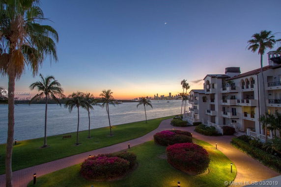 View of waterfront building with palm trees and skyline in the background.