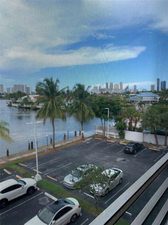 View of a parking area with several parked cars, palm trees, and buildings in the background.