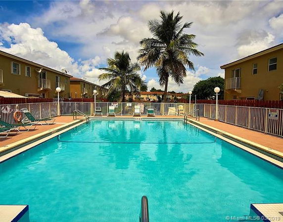 View of a swimming pool with adjacent buildings and palm trees in the background.