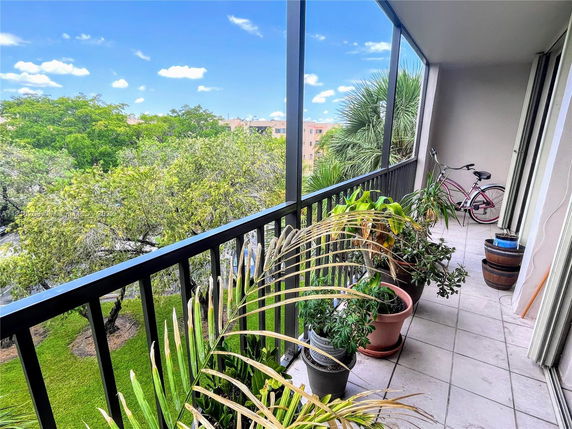 Balcony view with plants and a bicycle, overlooking green trees and a clear blue sky.
