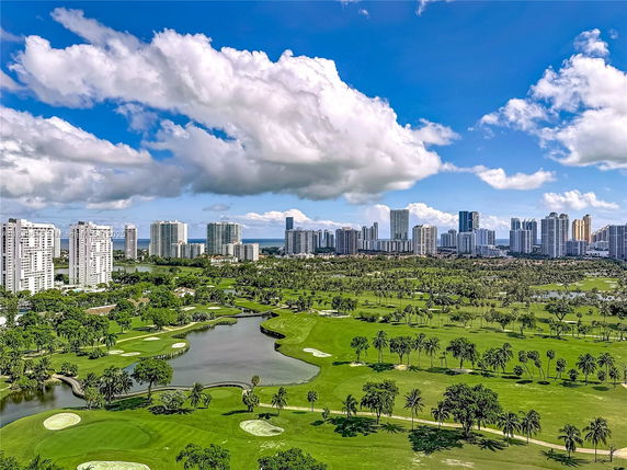 Panoramic view of a city skyline with numerous tall buildings, green golf course, and a cloudy sky.