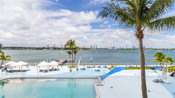 Panoramic view of a waterfront area with swimming pool, palm trees, and city skyline in the background.
