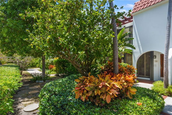 Front view of a house with arched doorway and windows, surrounded by lush greenery and garden plants.