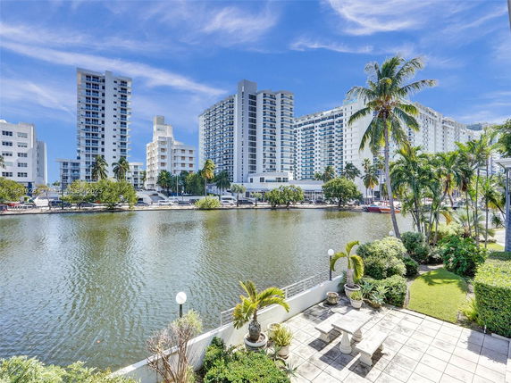 View of high-rise buildings across a waterway with palm trees and landscaping in the foreground.