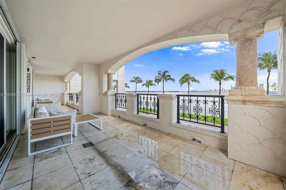 Covered patio area with seating, featuring ornate railings and arched columns, overlooking water and palm trees.