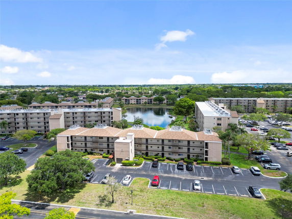 Aerial view of multiple apartment buildings with a parking area and greenery, surrounding a small lake.