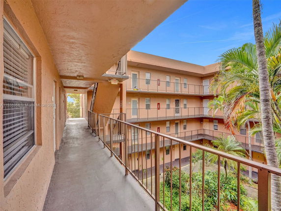 View of a multi-story building with open corridors and balustrades, surrounding a central courtyard with palm trees and greenery.