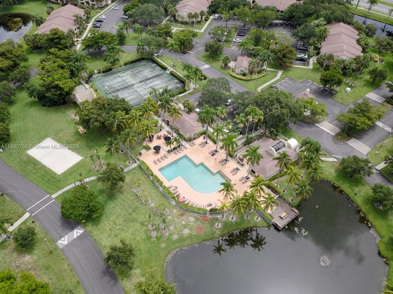 Aerial view of a residential area with a large swimming pool, tennis courts, parking areas, and surrounding greenery.