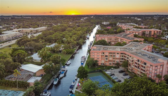 Aerial view of residential buildings along a canal with boats and a tennis court visible. Sunset in the background.