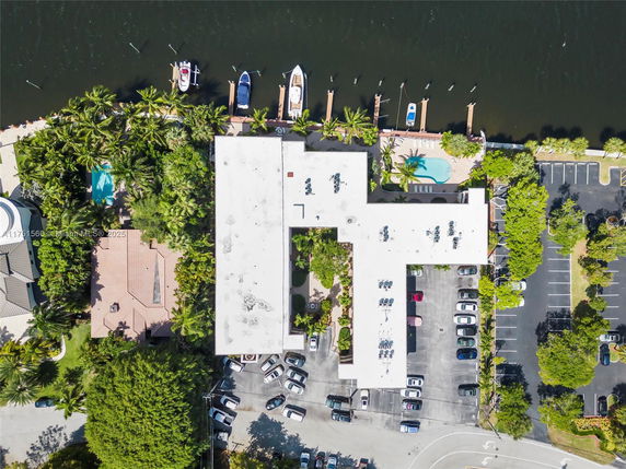 Aerial view of a building along a waterfront with a white rooftop, parking area, and a nearby pool.