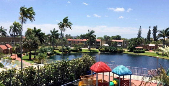 Scenic view of residential buildings, palm trees, and a pond, with a glimpse of a play area with colorful roofs.