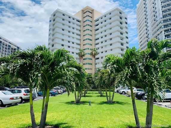 View of tall residential buildings with balconies, surrounded by palm trees and parked cars.