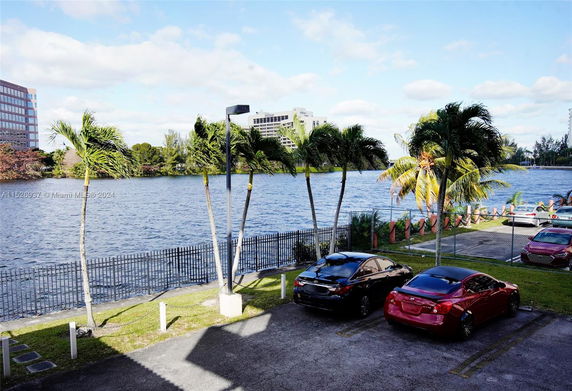 Outdoor parking area with cars, near a waterfront, and palm trees in view.