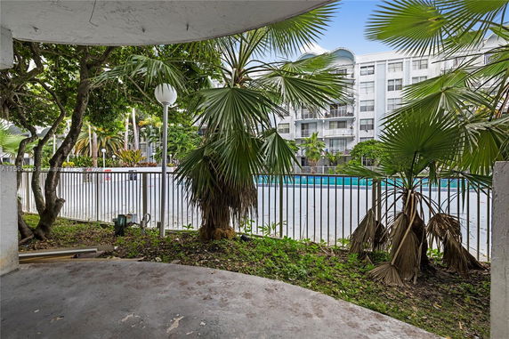 View of an outdoor swimming pool area with surrounding trees and a multi-story building in the background.