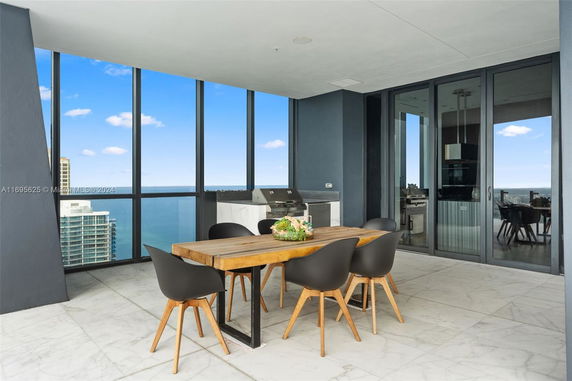 Balcony view from a high-rise building featuring a wooden dining table, modern chairs, and a built-in grill area with a view of the ocean and neighboring buildings.