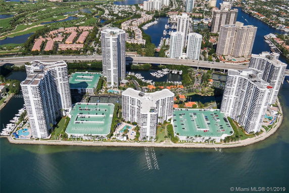 Aerial view of a waterfront high-rise complex with multiple white buildings and green-roofed parking structures.