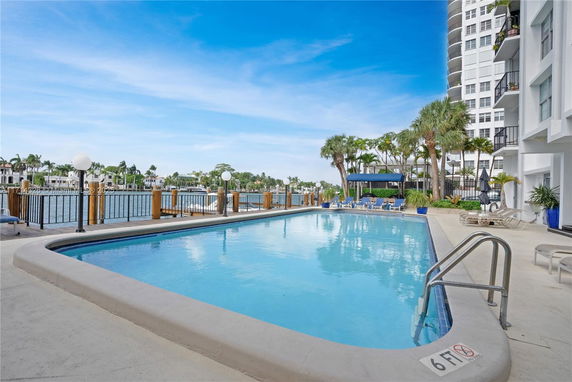Outdoor swimming pool area adjacent to a waterfront with a high-rise building.
