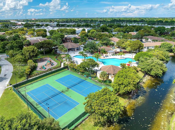 Aerial view of a community area with tennis courts, a playground, swimming pools, and surrounding greenery.