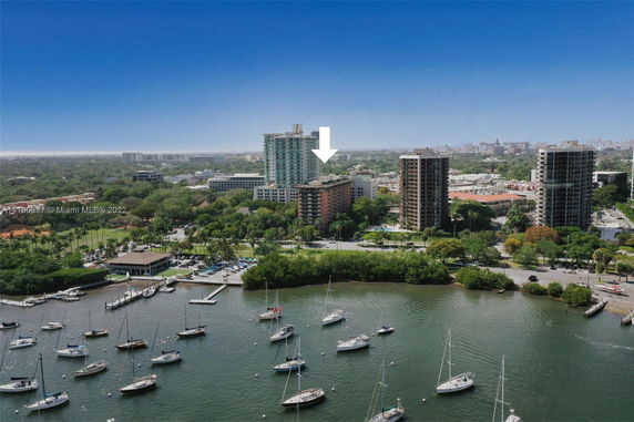 Aerial view of a high-rise residential building highlighted by an arrow, surrounded by other buildings, greenery, and a marina with boats.