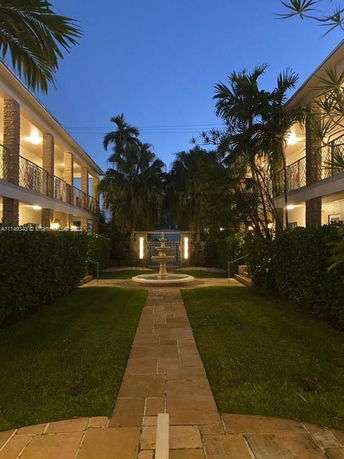 Pathway between two-story buildings with evening lighting and a central fountain.