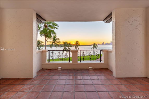 Terrace with tiled floor and ornate railing, offering a view of the ocean, palm trees, and a distant city skyline at sunset.