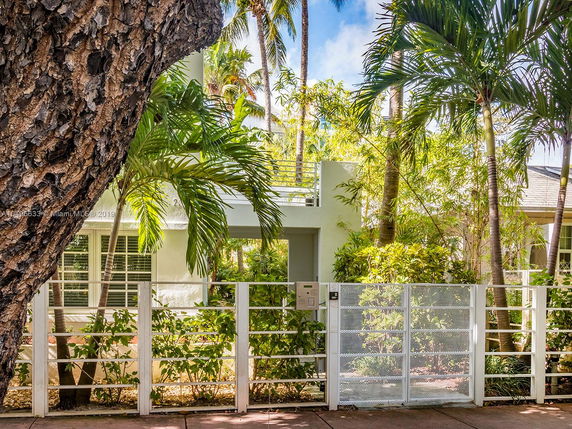 Front view of a house with a white fence and surrounded by palm trees.