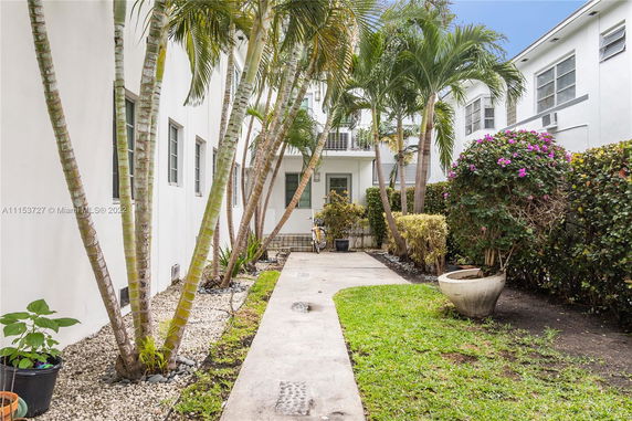 Pathway alongside white residential building with palm trees and shrubs.